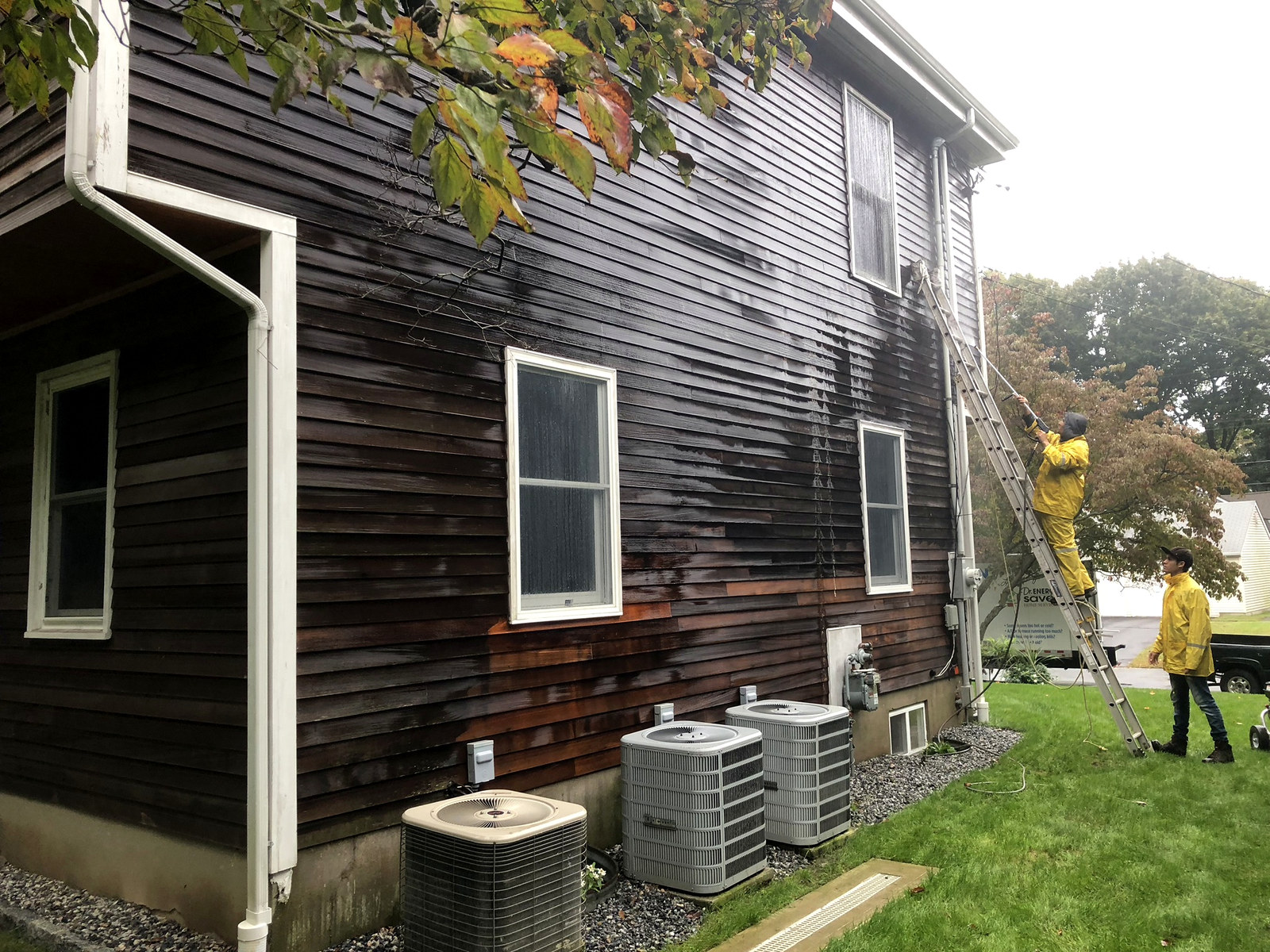 Crew in branded yellow rain gear pressure-washing dark cedar exterior